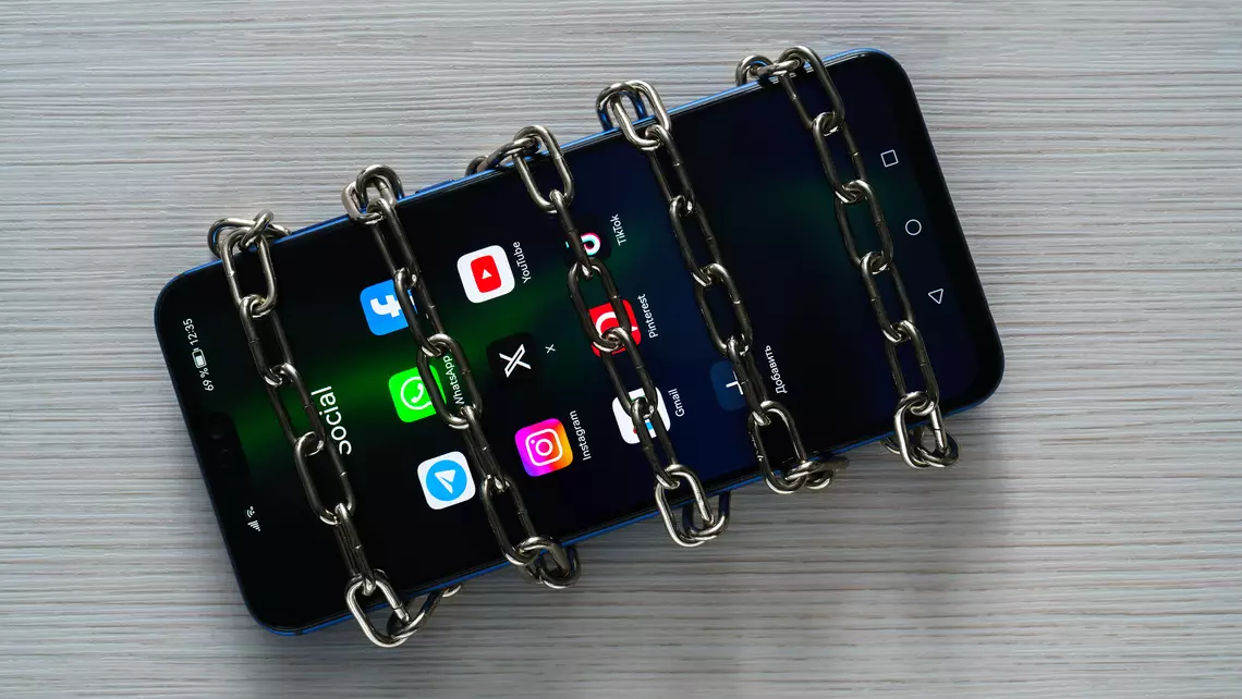 Smartphone avec différentes icônes de réseaux sociaux sur l'écrans et des chaînes autour du téléphone signifiant l'interdiction de ces réseaux