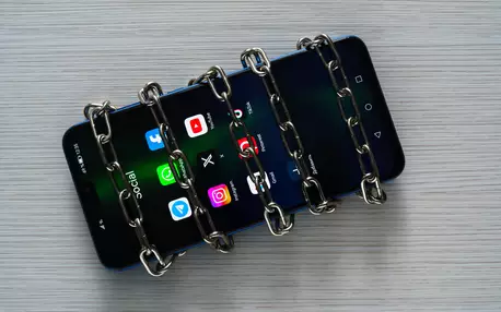 Smartphone avec différentes icônes de réseaux sociaux sur l'écrans et des chaînes autour du téléphone signifiant l'interdiction de ces réseaux